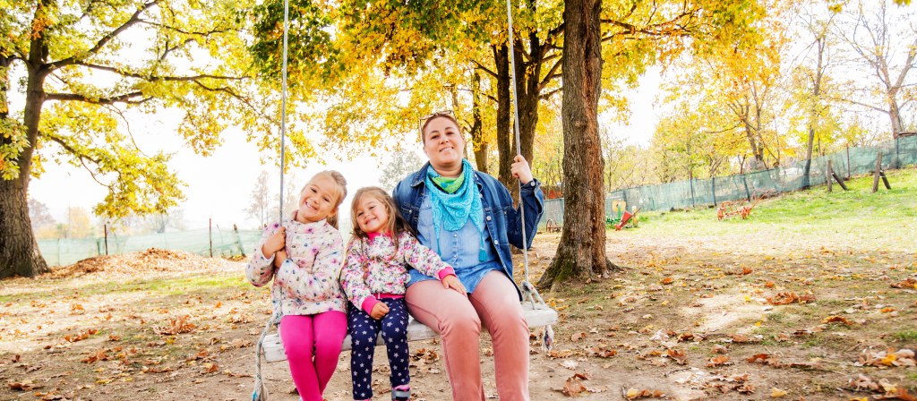 Happy Family Relaxing On Swing In Park. Smiling mother with daughters fun on sunny autumn day in nature.